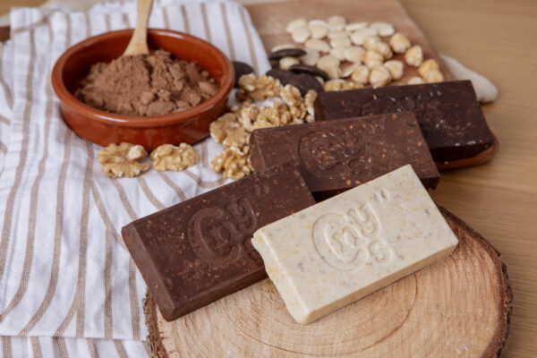 Variedad de barras de turrón artesanal de Chocolates Canayas en chocolate negro, con leche y blanco, presentadas sobre una base de madera junto a nueces, almendras y cacao en polvo.